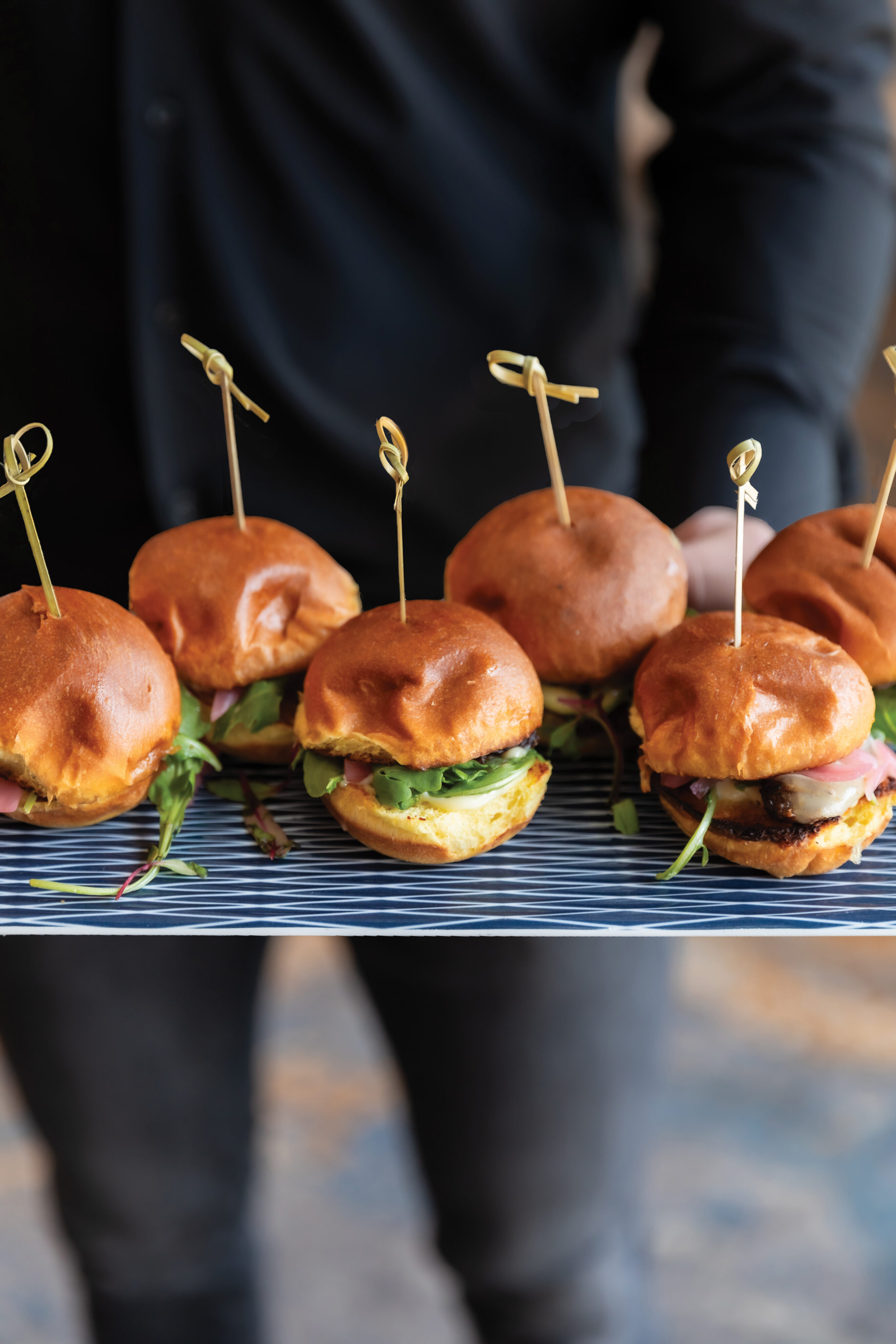 A person holds a tray of six mini burgers with lettuce and skewers, served on a striped platter at the lively Golf Suite.