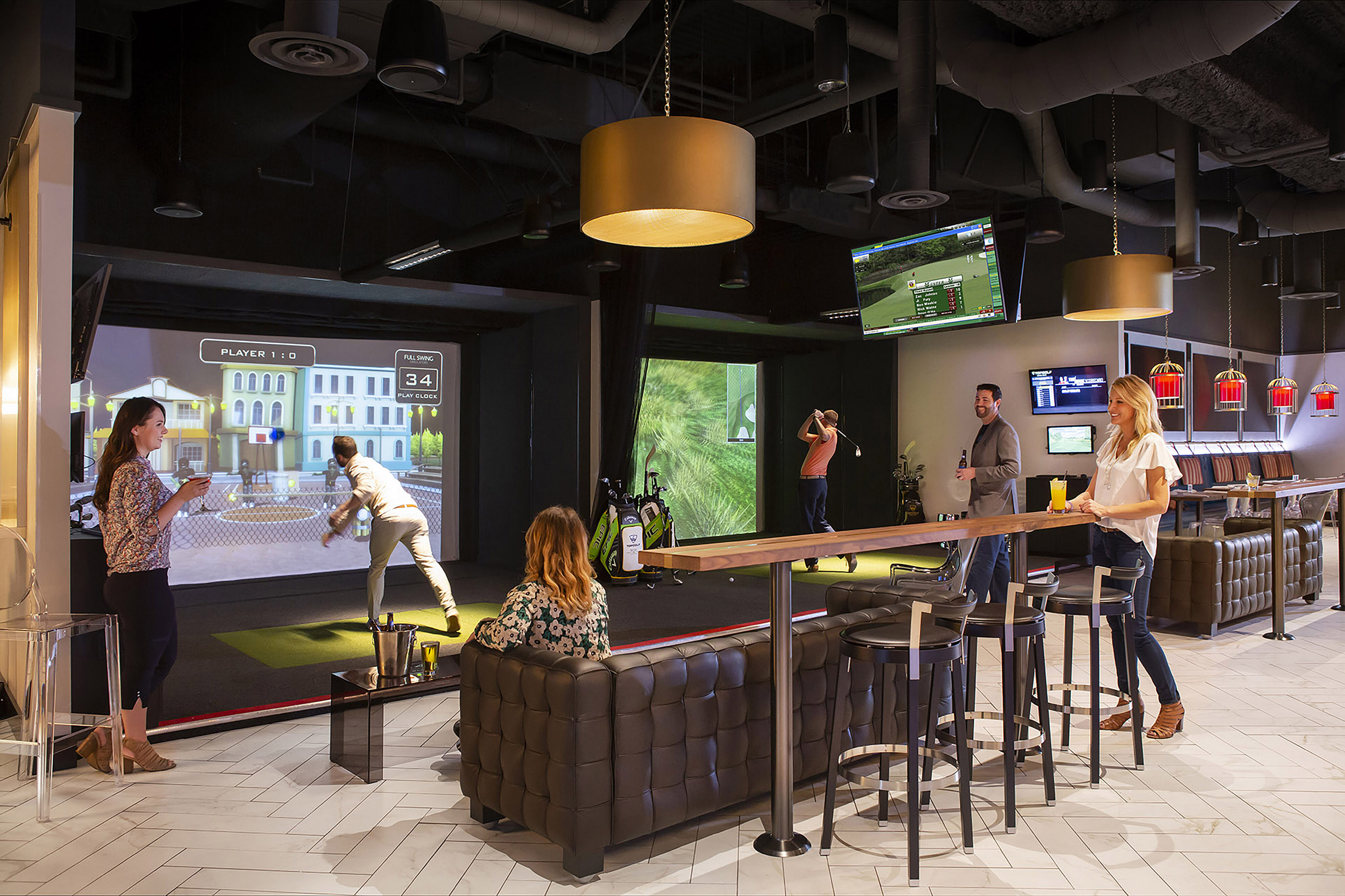 People play virtual golf in the Tempo Top Golf Suite, an indoor entertainment venue with seating, screens, and a bar area; others watch and interact nearby.