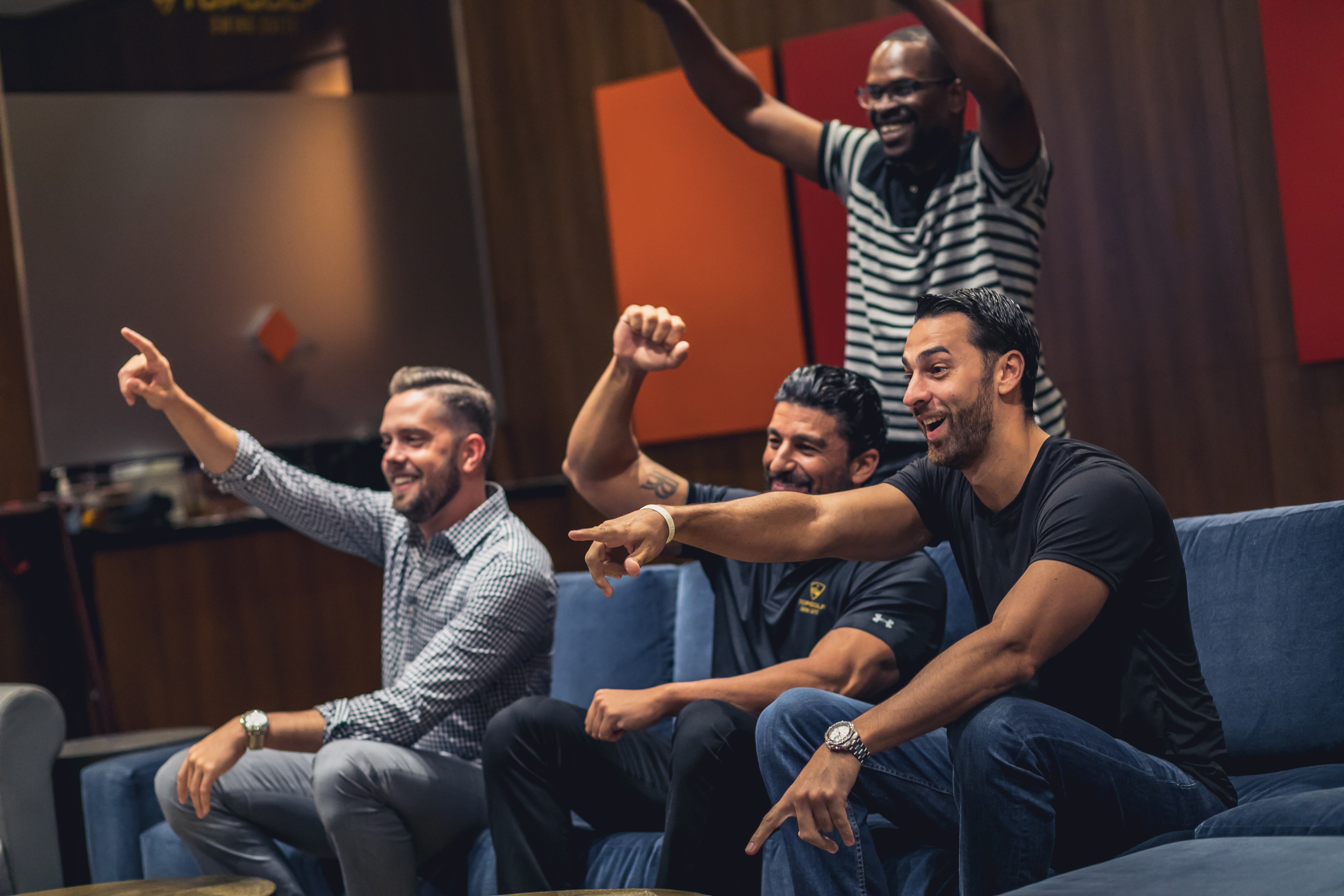 Four men sitting and standing in a living room, smiling and pointing excitedly at something off-camera, appearing engaged and animated as they enjoy a Tempo Top Golf Suite experience.